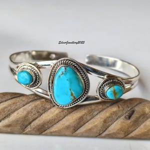 Könnte beinhalten: Silbernes Armreif-Armband mit drei Türkissteinen. Der zentrale Stein ist oval, die seitlichen Steine sind rund. Das Armband hat ein geflochtenes Seildesign um die Steine. Der Text "SilverJewellery2025" ist sichtbar. Auf einer Holzoberfläche.