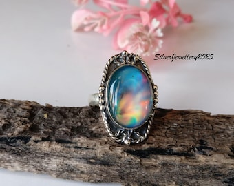 May include: Oval-shaped silver ring with a large, iridescent gemstone. The gemstone displays a blend of blue, orange, and yellow hues. The ring has a detailed, rope-like border and the text "SilverJewellery2025".