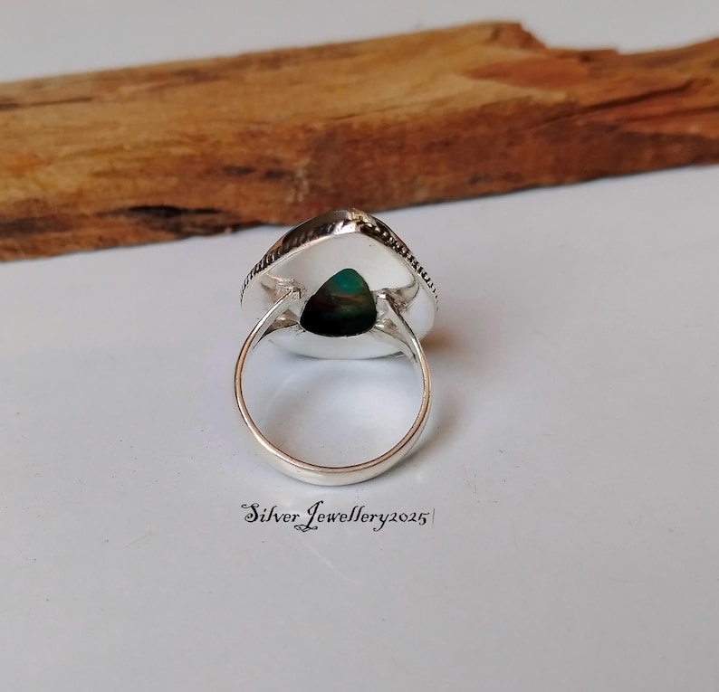 May include: A silver ring with a green gemstone set in a triangular shape. The ring is on a wooden surface.