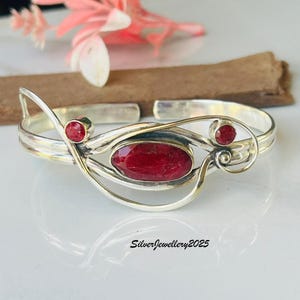 May include: A silver cuff bracelet with three red gemstones. The central gemstone is oval-shaped, and the two smaller gemstones are round. The bracelet has a flowing, open design. The text "SilverJewellery2025" is at the bottom.