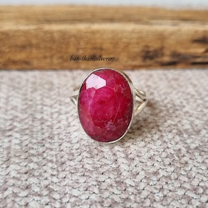 May include: A silver ring featuring a large, oval-shaped, faceted ruby gemstone. The ring has a simple, elegant design.