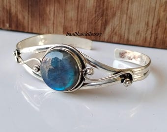 Puede incluir: Un brazalete de plata con un gran gemas de labradorita azul redonda engastada en el centro. El brazalete tiene un diseño delicado y curvo con pequeñas cuentas de plata.