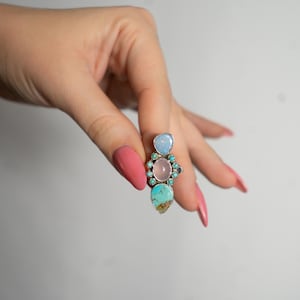 May include: A silver ring with a cluster of colorful gemstones. The ring features a light blue oval stone at the top, a pink round stone in the center, and a turquoise teardrop-shaped stone at the bottom. Small blue stones surround the pink stone.