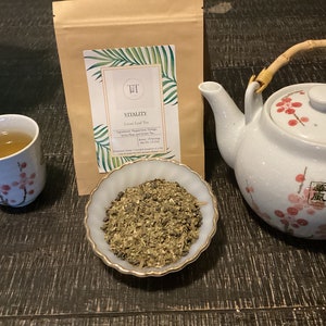 May include: A white teapot with a bamboo handle and pink floral design, a small white cup with pink floral design filled with green tea, a small white bowl with loose leaf tea, and a brown paper bag with a label that reads "Vitality Loose Leaf Tea" with a green palm leaf design.
