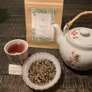 May include: A tea set featuring a white teapot with a bamboo handle and floral design, a teacup with tea, a small dish of loose leaf tea, and a bag of tea labeled "Hiblicious". The tea is a reddish-pink color.