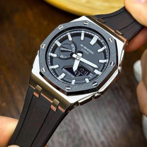 May include: A G-Shock Casio watch with a black dial, silver and black bezel, and a black rubber strap. The watch displays the time as 17:13. The watch has a silver case and a black band.