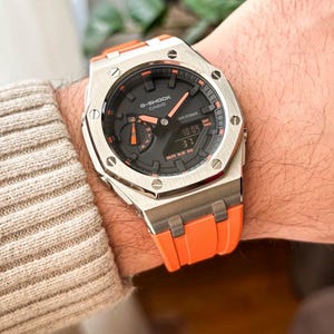 Op de afbeelding: Een zilverkleurig G-Shock Casio horloge met een zwarte wijzerplaat en oranje accenten. Het horloge heeft een digitale display met de tijd. De horlogeband is oranje. Het horloge zit om een pols.