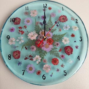 May include: Round wall clock with a light blue face, featuring dried flowers in shades of red, pink, purple, and white. Black numerals mark the hours. The clock hands are black and red.