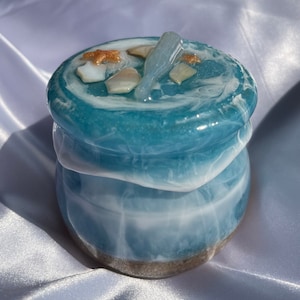 May include: A small, round, blue and white resin jar with a lid. The jar is decorated with seashells and a starfish. The jar has a sandy bottom.