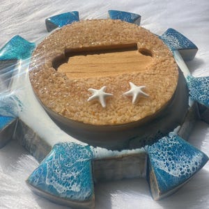May include: A decorative resin tray with a beach theme. The round center has a sandy texture with two white starfish. The tray is surrounded by blue and white resin accents, resembling ocean waves. The tray is approximately 10 inches in diameter.