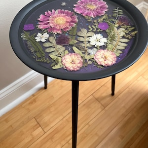 May include: A round, black side table with a floral design. The tabletop features pressed flowers in shades of pink, purple, and white, set against a dark background. The table has three slender black legs.