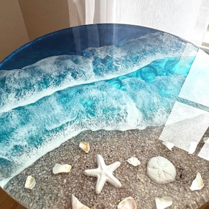May include: Round resin table with a beach scene design. The table features a clear top with a blue ocean and white foamy waves. The beach area includes a white starfish, seashells, and a sand dollar.