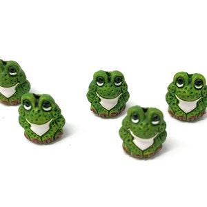 May include: Five green frog buttons with white bellies and red feet. The frogs have black eyes and are sitting on a white surface.