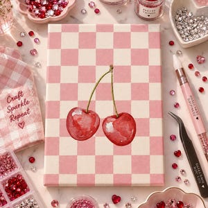 May include: A rectangular canvas featuring a pink and white checkered pattern with a central watercolor-style illustration of two red cherries. The canvas is surrounded by various containers of red, pink, and clear rhinestones, a pink stylus, and tweezers.