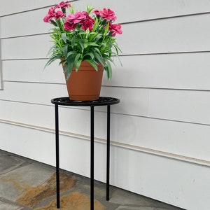 May include: A black metal plant stand with a round top and three legs. A terracotta pot with pink flowers sits on the stand.