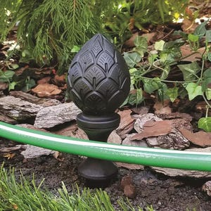 May include: A matte black garden stake featuring a finial shaped like a stylized pinecone or artichoke with layered, textured scales. The decorative topper sits atop a tapered, smooth pedestal base that is anchored directly into the dark soil.