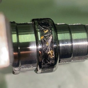 以下が含まれることがあります： 金属旋盤で加工されている黒と金のリングのクローズアップ。リングは、金色のフレークが埋め込まれた黒いカーボンファイバーのバンドを持っています。