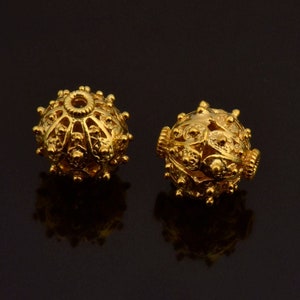 May include: Two gold-colored metal beads with intricate, detailed designs. The beads are round and have a hole in the center for stringing.
