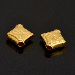 May include: Two gold-coloured square-shaped beads with a decorative floral design. The beads have a smooth, polished finish.
