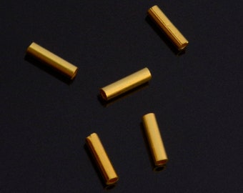 May include: Six gold-colored metal tubes, likely used for jewelry making, are scattered on a black surface.