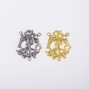 以下が含まれることがあります： 翼のある人物が剣を持っている金属製のペンダントが2つ。1つはシルバーカラー、もう1つはゴールドカラーです。各ペンダントには、ジュエリーやその他の工芸品に取り付けるための小さな穴が2つあります。ペンダントは白い背景にあります。