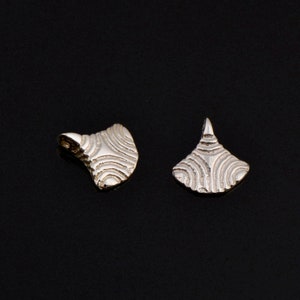 Könnte beinhalten: Zwei silberfarbene Metallanhänger mit geometrischem Design. Die Anhänger sind fächerförmig und haben eine strukturierte Oberfläche.
