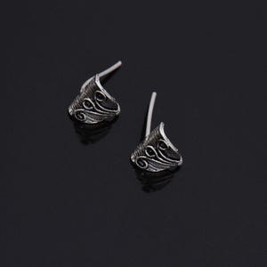May include: A pair of silver stud earrings. Each earring features a triangular design with a curved top and decorative swirl patterns. The posts are white. The earrings are on a black surface.