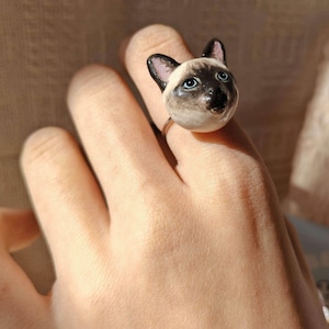 Könnte beinhalten: Ein Ring mit einem Siamkatzenkopf-Design. Das Gesicht der Katze ist mit schwarzen, braunen und weißen Farben, blauen Augen und schwarzen Ohren detailliert. Der Ring ist aus dünnem, goldfarbenem Metall. Der Katzenkopf ist das Hauptmerkmal des Rings.