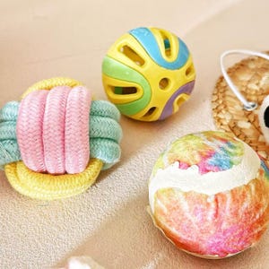 May include: A collection of colorful pet toys. Includes a yellow and blue ball with cutouts, a multi-colored tie-dye ball, and a pink, yellow, and green rope toy. These toys are designed for pet play and enrichment.