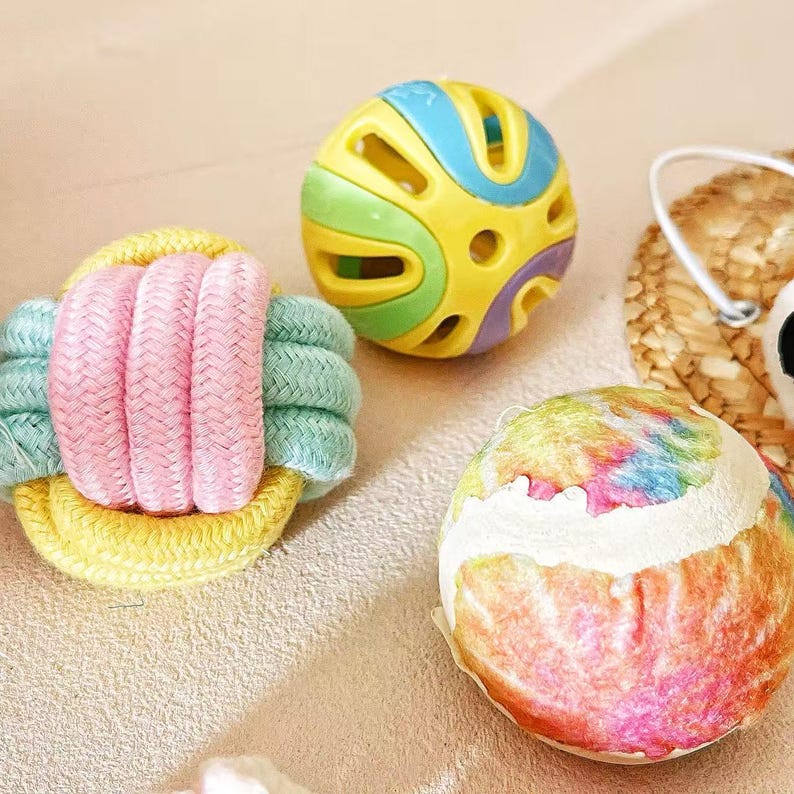 May include: A collection of colorful pet toys. Includes a yellow and blue ball with cutouts, a multi-colored tie-dye ball, and a pink, yellow, and green rope toy. These toys are designed for pet play and enrichment.