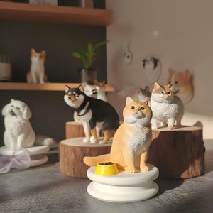 Puede incluir: Una colección de figuras de cerámica de animales, incluyendo gatos y perros, en varias poses. Las figuras son en tonos de blanco, negro y naranja, y algunas están sobre plataformas de madera. Un cuenco amarillo está delante de un gato.
