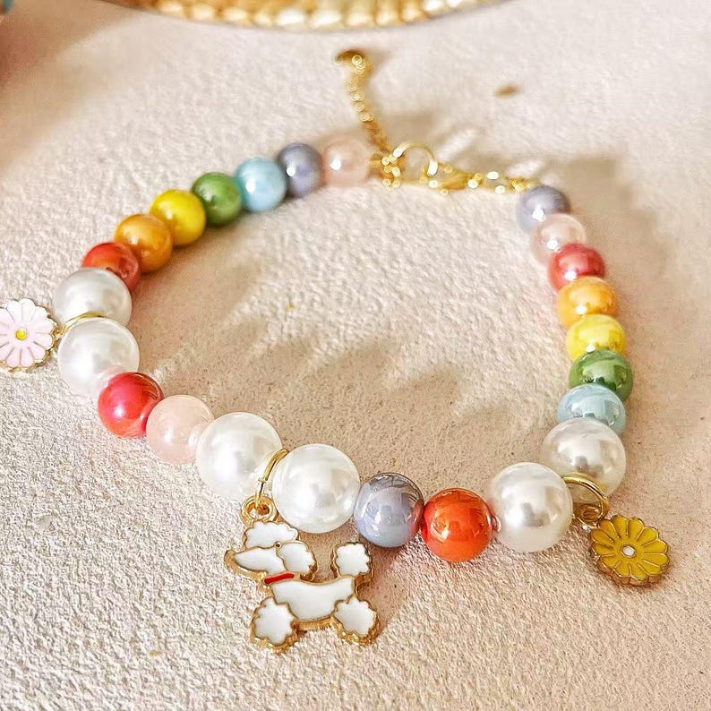 May include: A colorful beaded bracelet featuring a variety of beads in rainbow hues and large white faux pearls. The bracelet includes gold charms of a poodle and a yellow flower. The bracelet is on a light pink surface.