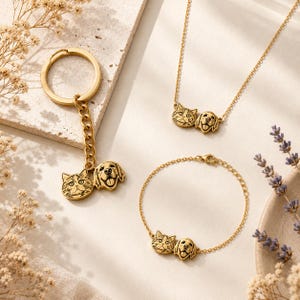 May include: Gold-colored jewelry set featuring a cat and dog design. The set includes a keychain, necklace, and bracelet. The cat and dog pendants are detailed with facial features. The jewelry is displayed on a neutral surface with floral accents.