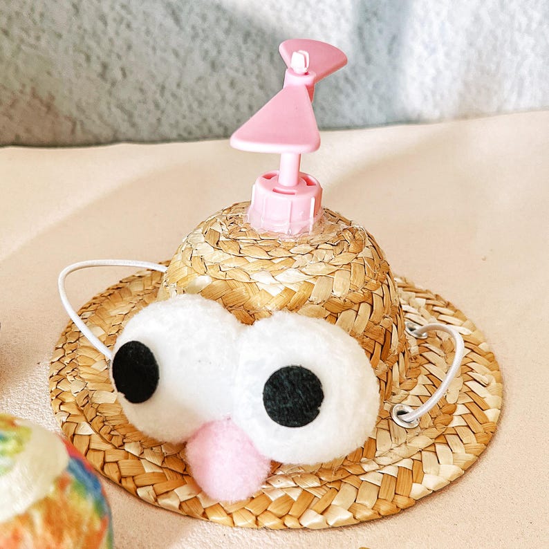 May include: A novelty pet hat with a straw brim, large white plush eyes with black pupils, and a pink nose. A pink plastic piece is attached to the top of the hat. A white elastic band is attached to the hat.
