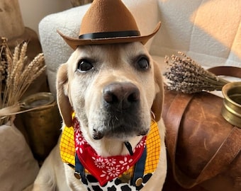 May include: A light-colored dog wearing a brown felt cowboy hat with a black band and a colorful bandana featuring a red floral pattern, a yellow plaid section, and a black and white cow print section with small gold-colored circular accents.