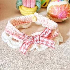 May include: A handmade crochet pet collar in white and pink, featuring a scalloped edge and a bow with a red and white pattern. The collar has a button closure. The background includes decorative Easter eggs.