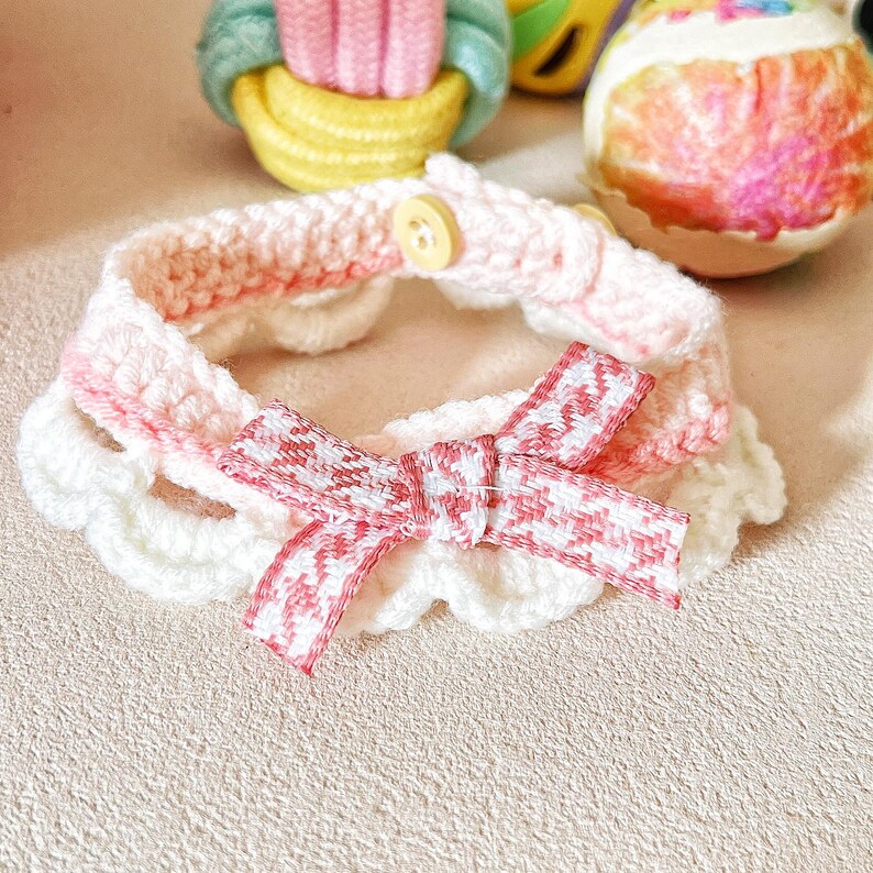 May include: A handmade crochet pet collar in white and pink, featuring a scalloped edge and a bow with a red and white pattern. The collar has a button closure. The background includes decorative Easter eggs.