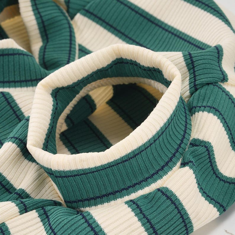 Pu&ograve; includere: Primo piano di un maglione a righe verdi e crema. Il tessuto a maglia a coste presenta ampie righe verdi con sottili accenti blu navy, alternate a righe color crema. Il colletto e i polsini sono color crema.