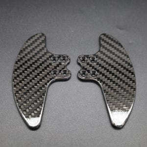 May include: Two carbon fiber paddle shifters, featuring a woven black and gray pattern. The shifters have a curved design with multiple mounting holes and a silver finish on the bottom edges. They are set against a gray background.