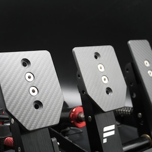 Può includere: Primo piano di un set di pedali per simulatore di corse. I pedali hanno una trama in fibra di carbonio e sono montati su un telaio nero. Il set include tre pedali, ciascuno con un design distintivo e hardware di montaggio. L'estetica generale è elegante e moderna.