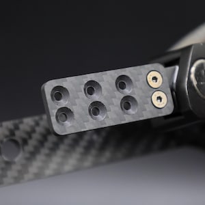 Puede incluir: Primer plano de un componente de fibra de carbono con múltiples orificios circulares y dos tornillos dorados. El componente forma parte de un conjunto más grande, de color oscuro, posiblemente automotriz o industrial.