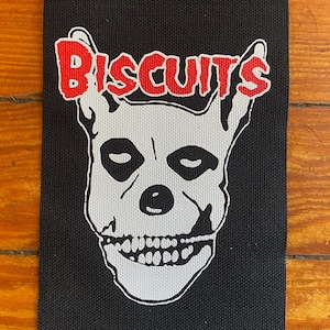 Pode incluir: Patch retangular preto com um desenho de caveira branca e a palavra "BISCUITS" em vermelho acima. A caveira tem órbitas oculares pretas e uma mandíbula detalhada. O patch é provavelmente para roupas ou acessórios.