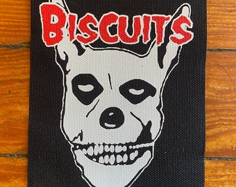 May include: Black rectangular patch featuring a white skull design with the word "BISCUITS" in red, stylized above the skull. The skull has black eye sockets and a detailed jawline. The patch is likely for clothing or accessories.