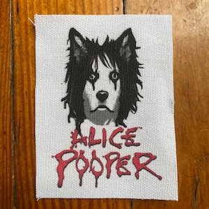 May include: A white fabric patch with a black and white illustration of a dog's head with a black and white striped face. The text "ALICE COOPER" is written in red below the dog's head.