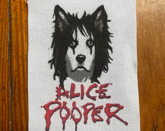 May include: A white fabric patch with a black and white illustration of a dog's head with a black and white striped face. The text "ALICE COOPER" is written in red below the dog's head.