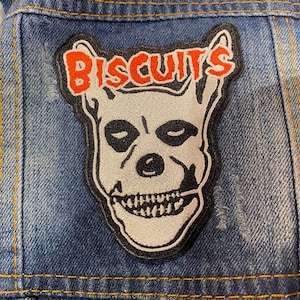Pode incluir: Uma caveira preta e branca com o texto "BISCUITS" em vermelho e branco acima dela. O remendo é costurado em uma jaqueta de jeans azul.