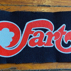 May include: A black fabric patch with a red and white stylized "fart" logo. The logo features a flower design.