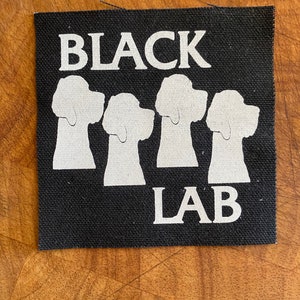 Puede incluir: Parche negro y blanco con el texto "BLACK LAB" y cuatro siluetas blancas de cabezas de Labrador Retriever.