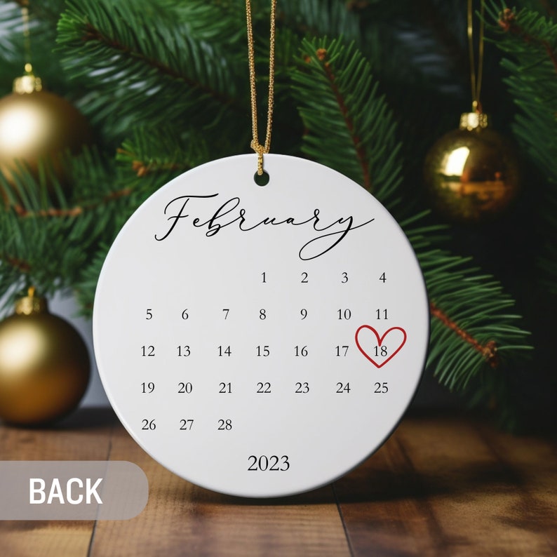May include: A round white ceramic ornament featuring the word February in black script, a calendar grid for the month, the year 2023, and a red heart outline encircling the number 18. The ornament hangs from a thin gold cord.