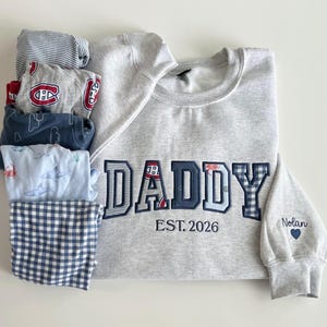 Peut inclure: Un sweat-shirt gris clair à col rond avec le mot DADDY brodé en lettres capitales dans des tissus à motifs variés, incluant un logo sportif, des rayures et des carreaux. Sous le mot, EST. 2026. La manche porte le nom Nolan et un petit cœur bleu.